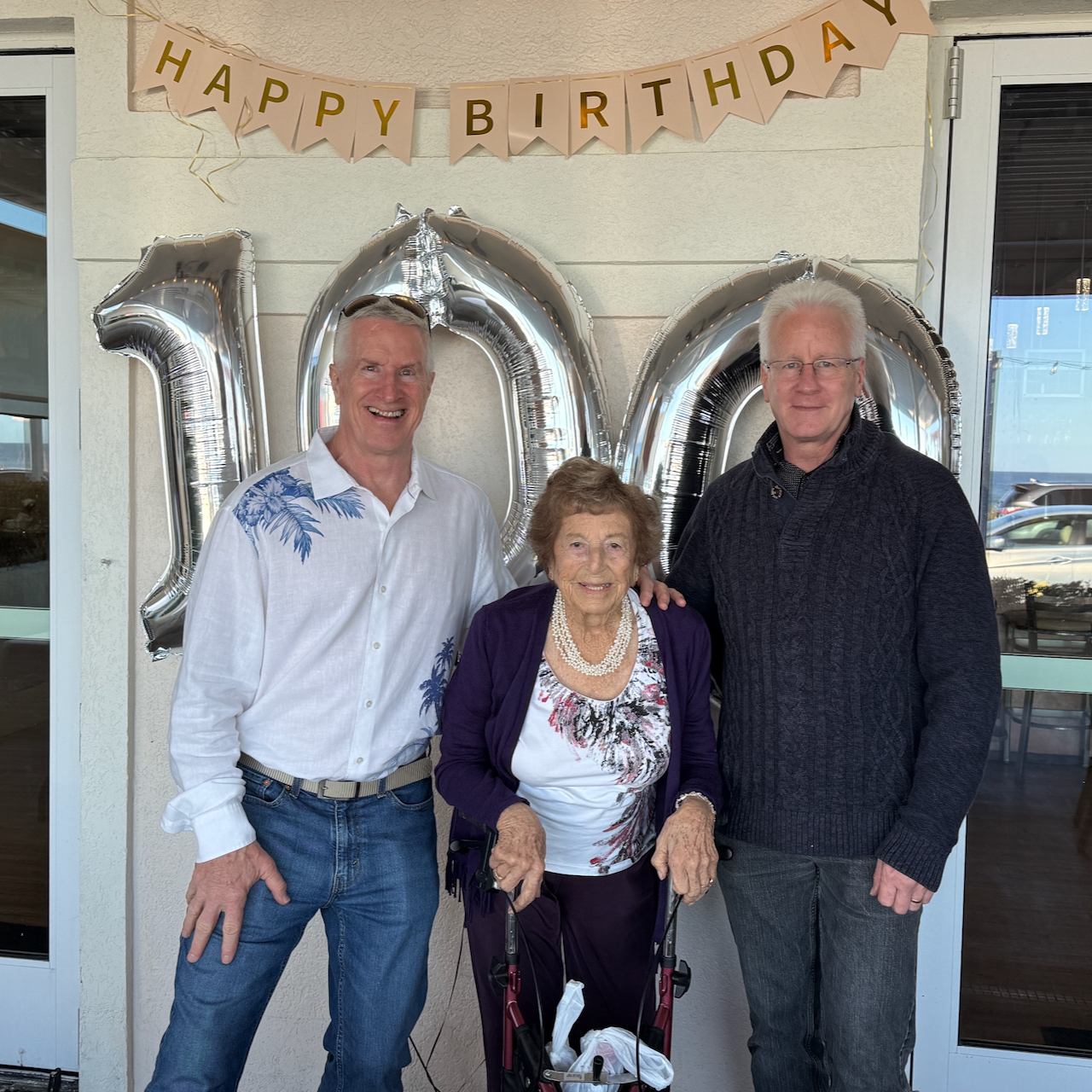 Colin and Paul Hammond with their mother Isobel outside Next Door Beach Bistro with Happy Birthday banner and silver 100 balloons