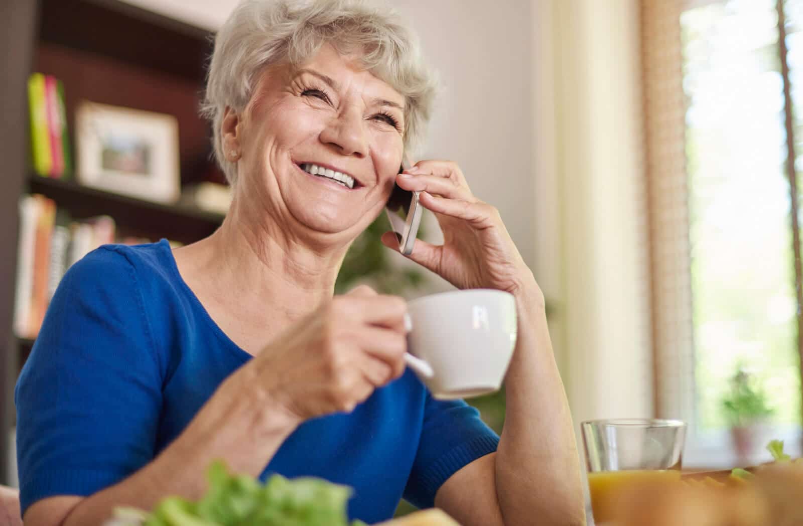 Happy senior woman smiling while talking on phone with coffee cup, demonstrating ease of daily check-in calls